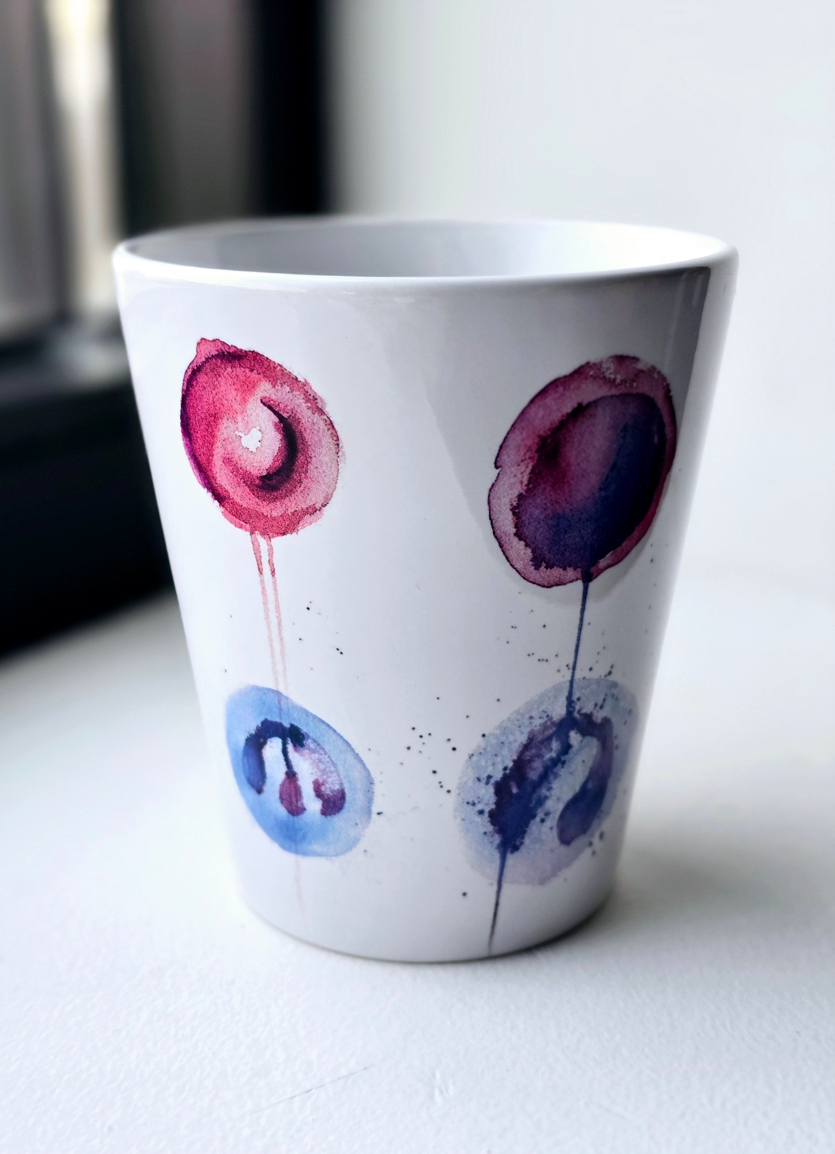 A white ceramic mug with a painting of six blood cells in blue, purple, and pink on a white background. Anatomy art gift for doctor or nurse.