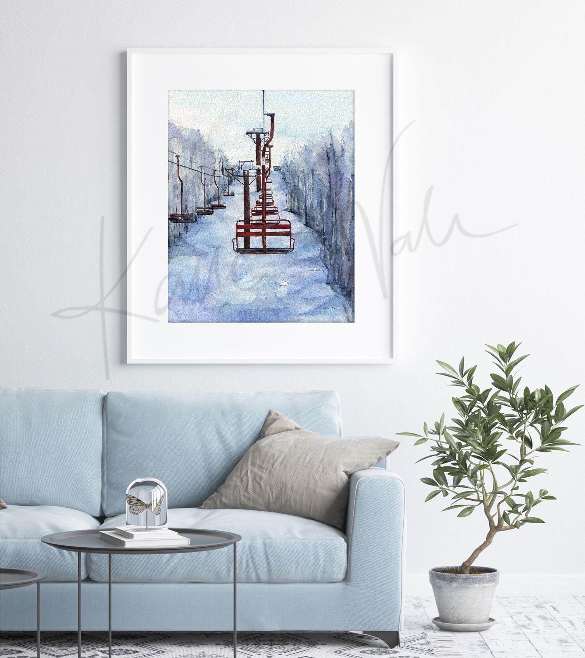 Framed watercolor painting of chairlifts in a snowy scene. The painting is hanging over a blue couch.