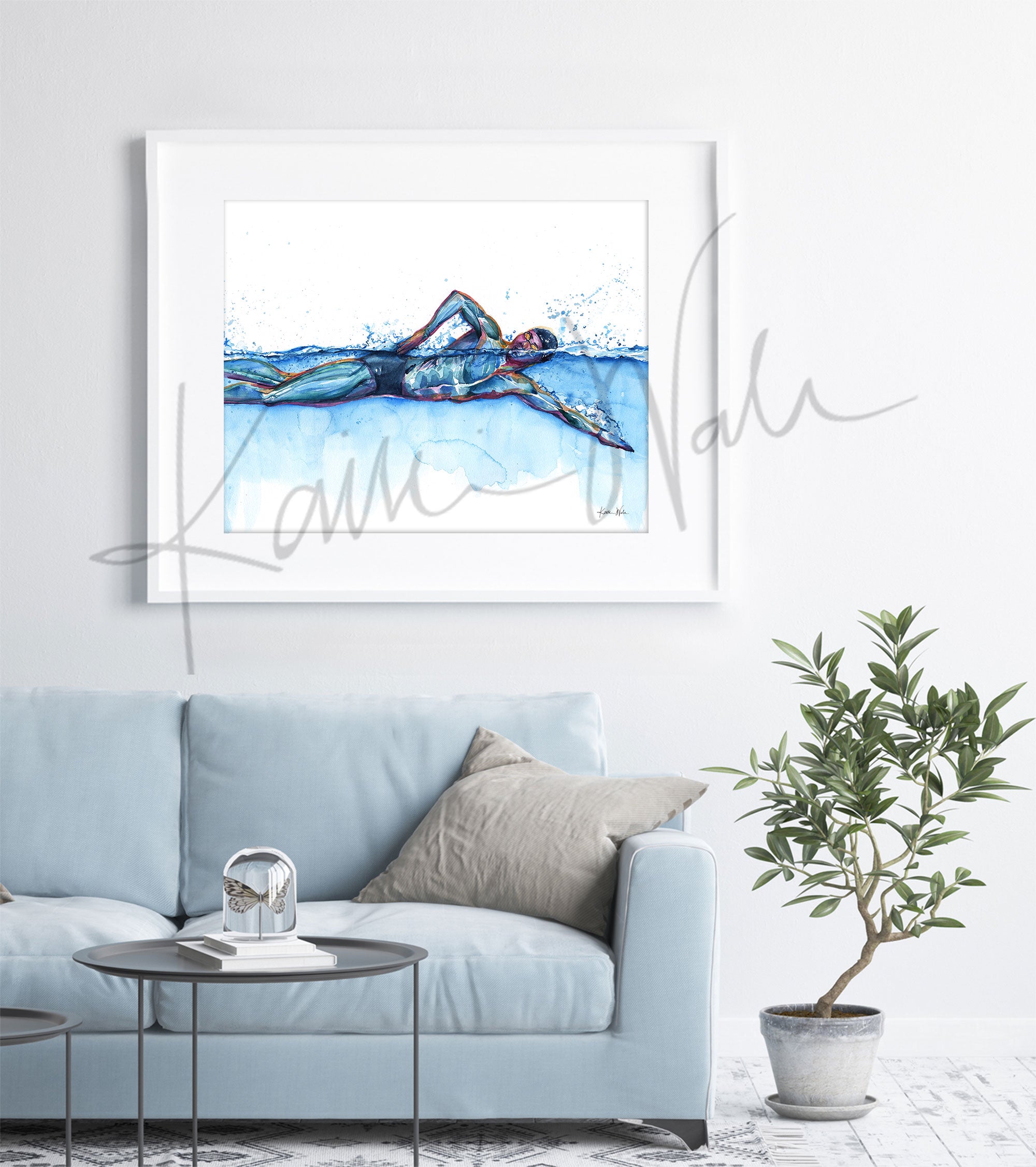 Framed watercolor painting of a swimmer in mid stroke showing the swimmer’s muscular anatomy. The painting is hanging over a blue couch.