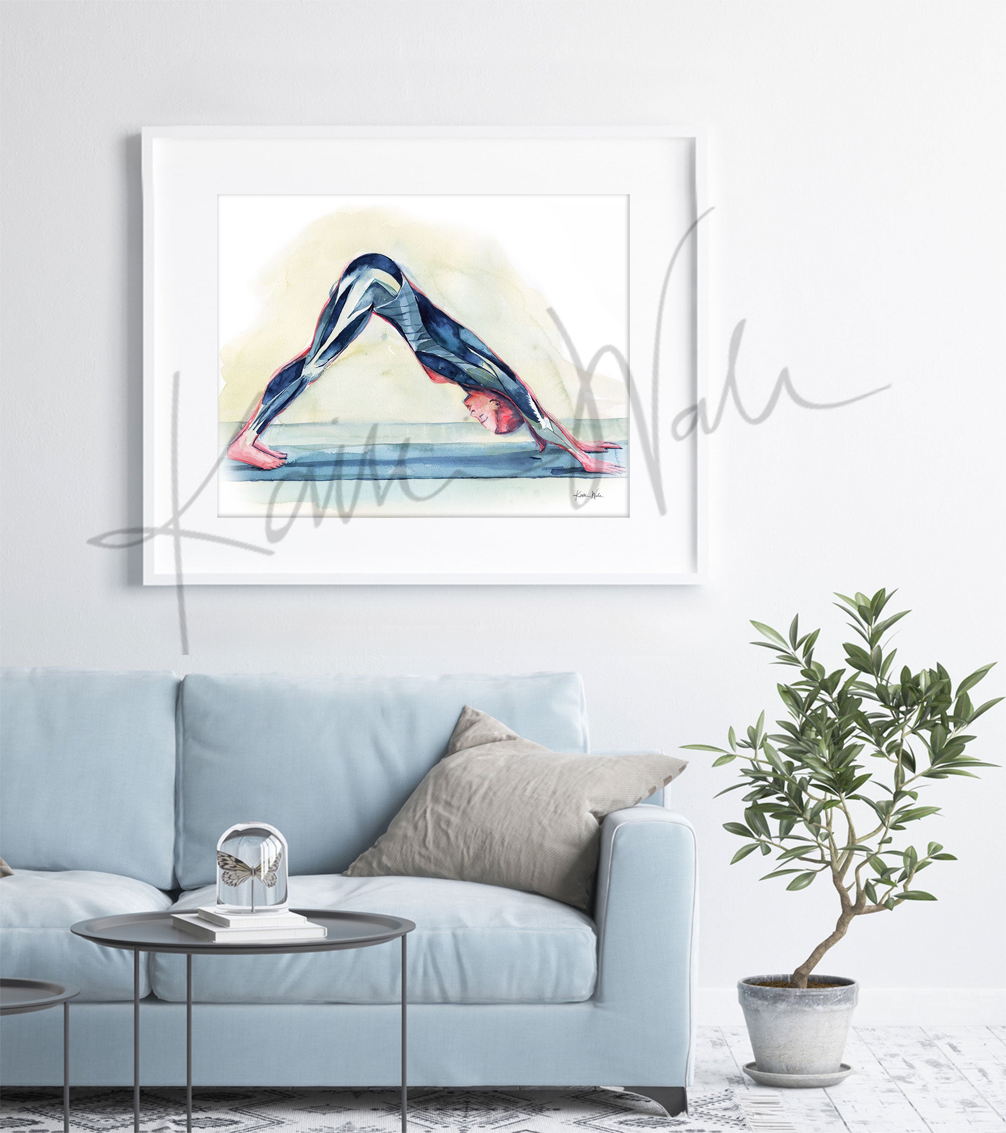 Framed watercolor painting of a woman doing a down dog yoga pose and shows her muscular anatomy within the pose. The painting is hanging over a blue couch.