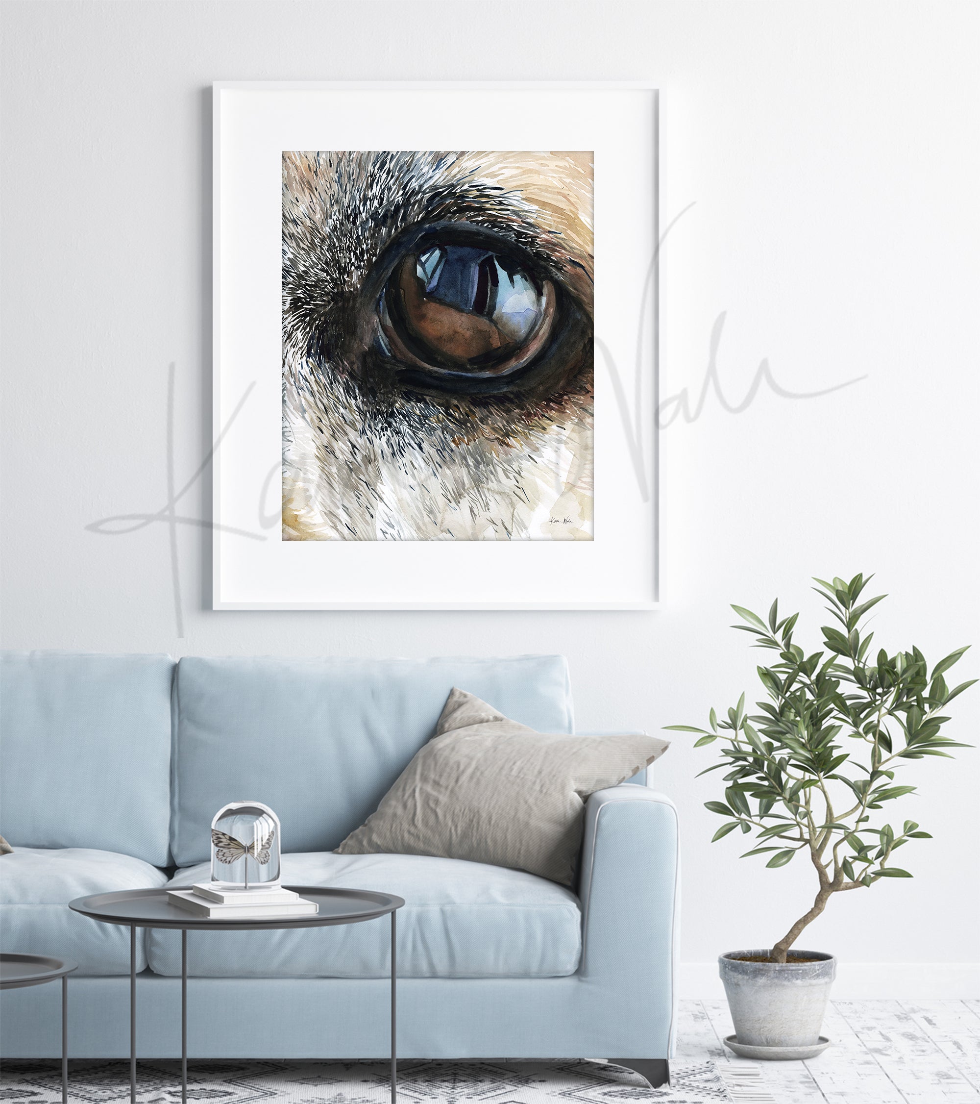 Framed watercolor painting of a zoomed in perspective of a dog’s eye. The painting is in blacks, tans, and browns. The painting is hanging over a blue couch. 