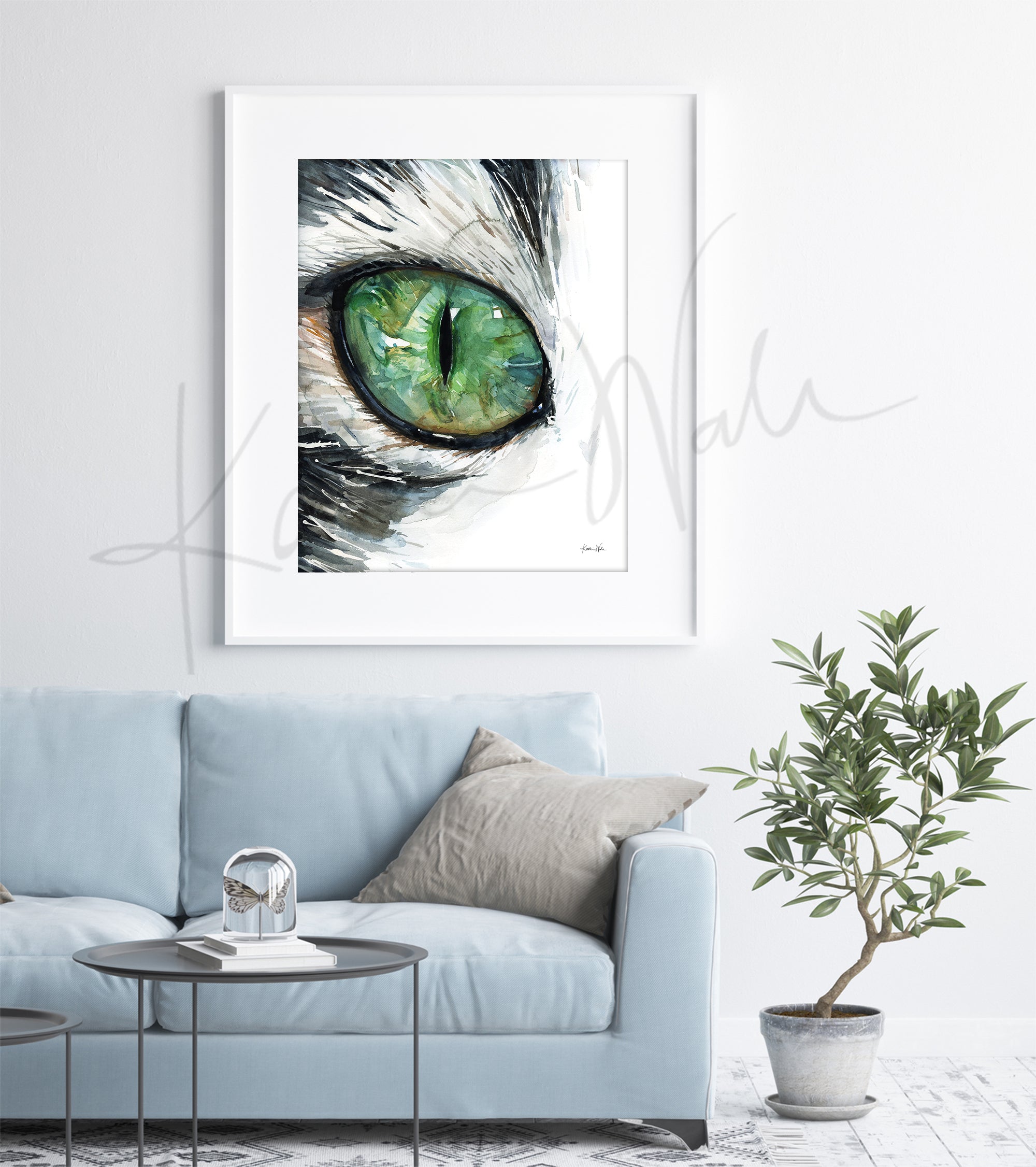 Framed watercolor painting of a zoomed in perspective of a green cat's eye. The hair surrounding the eye is black and white. The painting is hanging over a blue couch.