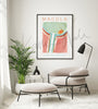 Framed contemporary poster design of the lens in teal, brown, orange, and gray. The painting is hanging over beige chair and footstool, plant, and black floor lamp.