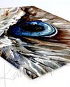 Unframed watercolor painting of a zoomed in perspective of an owl eye. The iris is blue and feathers around the eye are painted in tans and browns. The print is at an angle.