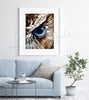 Framed watercolor painting of a zoomed in perspective of an owl eye. The iris is blue and feathers around the eye are painted in tans and browns. The painting is hanging over a blue couch.