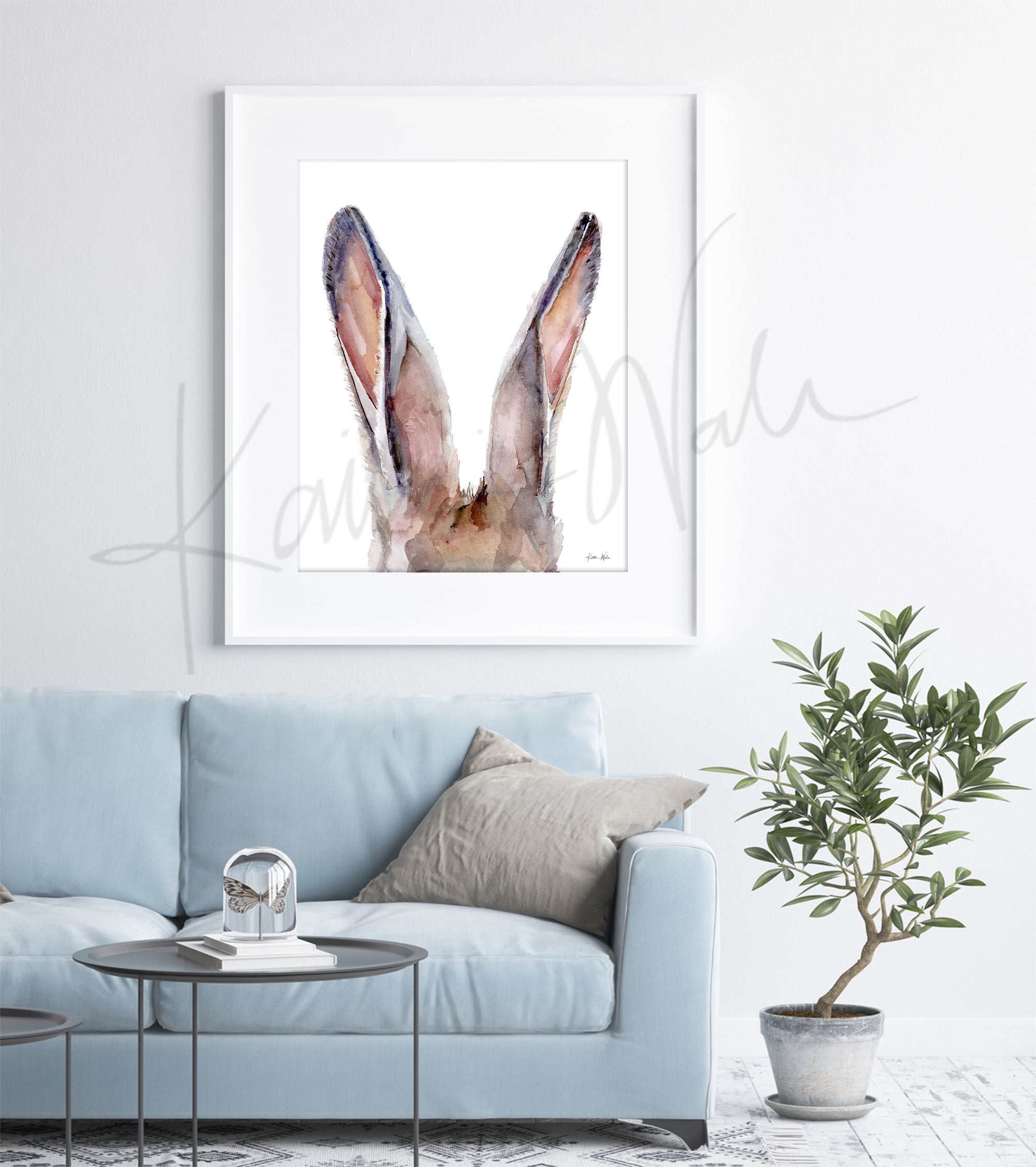 Framed watercolor painting of a zoomed in perspective of rabbit ears in tans, pinks, and browns. The painting is hanging over a blue couch.